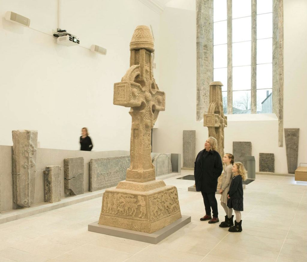 박물관에 커다란 석조 켈트 십자가 두 개가 있으며, 한 여성과 두 명의 어린이가 그들을 바라보고 있고 다른 한 사람이 배경에 있습니다.