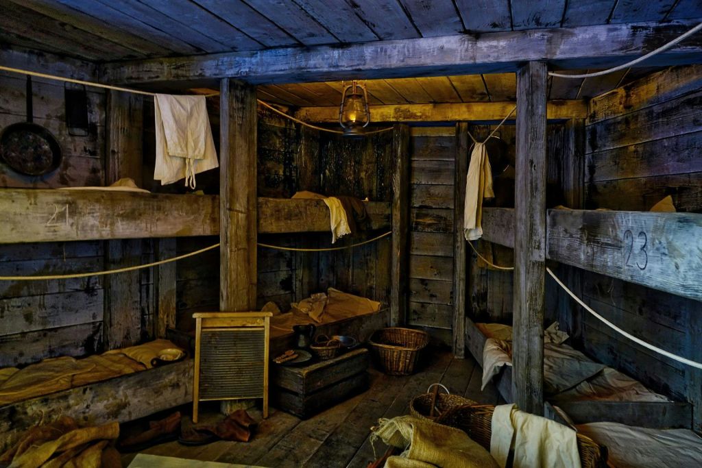 나무 이층 침대, 걸이식 세탁물, 랜턴, 세면대, 수납 바구니가 있는 조명이 희미하고 소박한 통나무집입니다.