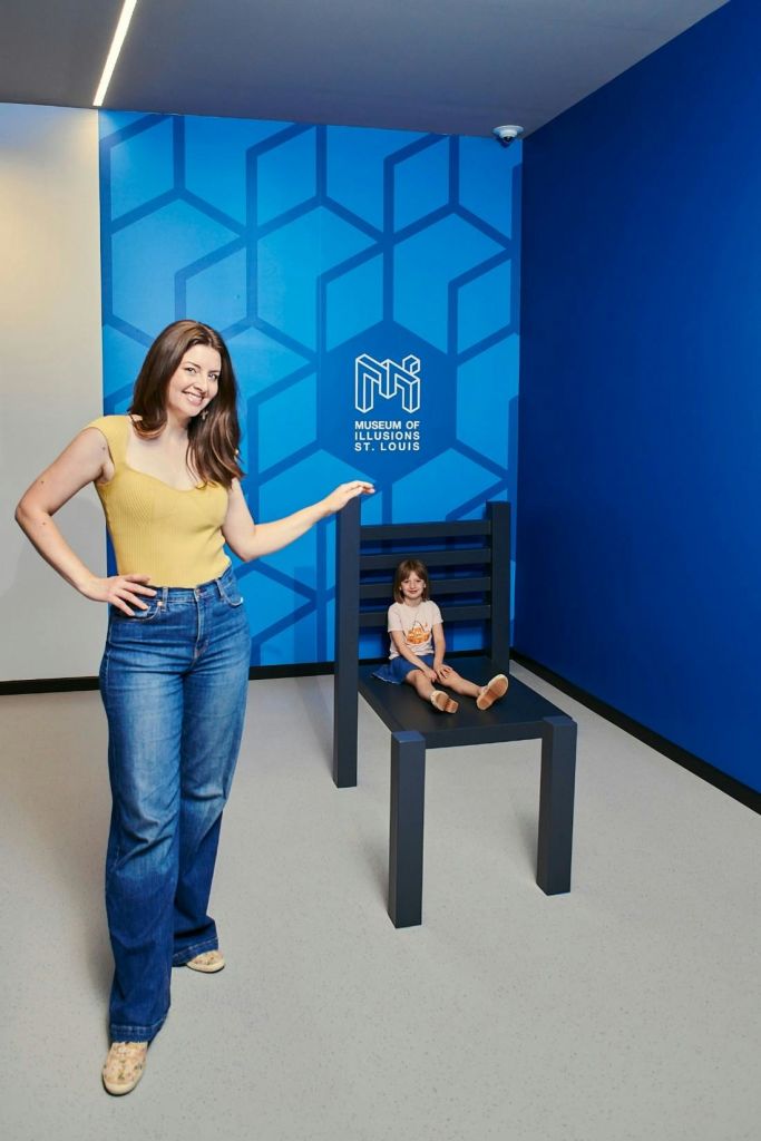한 여성이 아이가 앉아 있는 대형 의자 옆에 서서 미소를 짓고 있습니다. 파란색 벽에 Museum of Illusions St. Louis 로고가 있습니다.