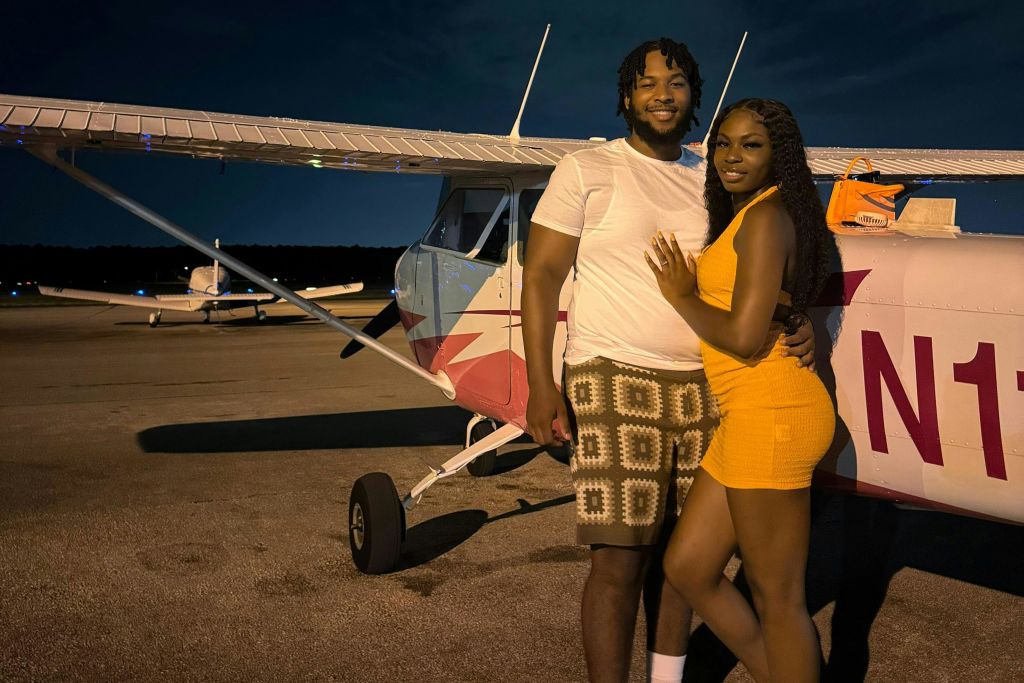 밤에 작은 흰색과 빨간색 비행기 앞에 미소 짓는 커플이 서 있습니다. 남자는 흰색 셔츠와 체크무늬 반바지를 입고, 여자는 오렌지색 드레스를 입습니다.