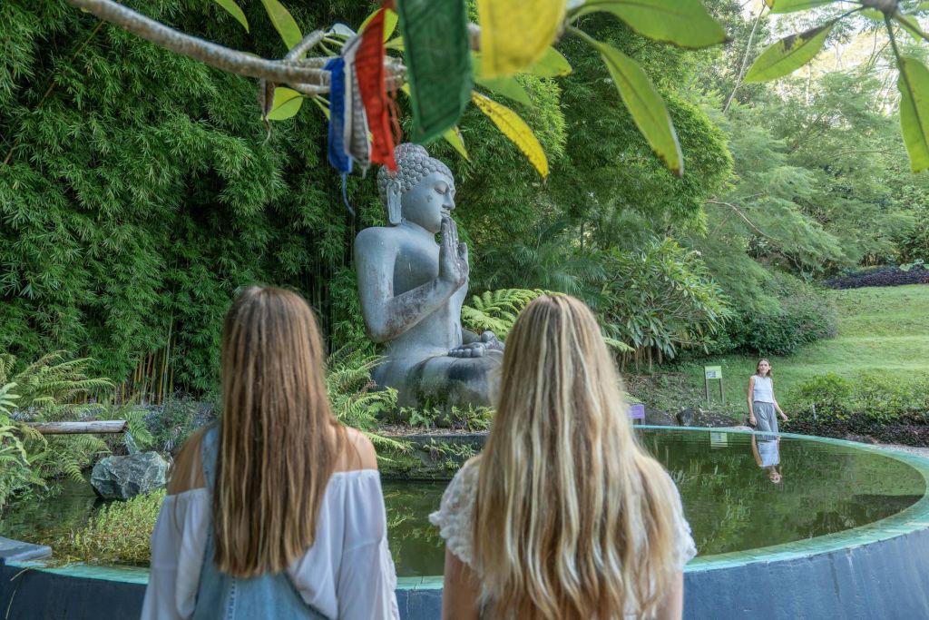 긴 머리를 한 두 여성이 울창한 녹음과 기도 깃발로 둘러싸인 연못 근처에서 석불상을 마주보고 서 있습니다.