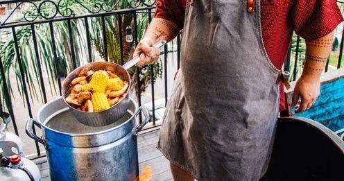 앞치마를 두른 사람이 옥수수와 새우가 담긴 냄비를 야외 파티오의 금속 용기 위에 올려놓고 있습니다.