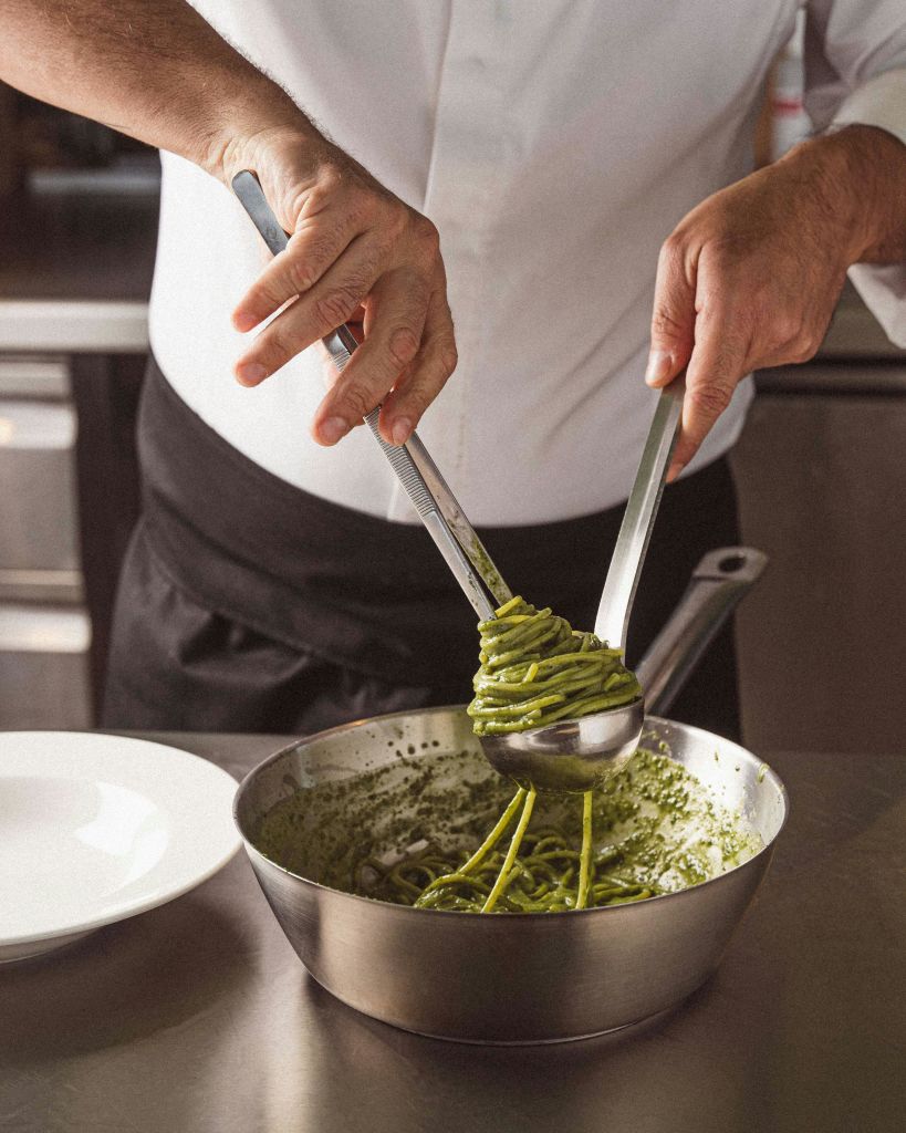 집게와 국자를 사용하여 팬에서 페스토 파스타를 제공하는 흰색 코트와 검은색 앞치마를 입은 셰프입니다.