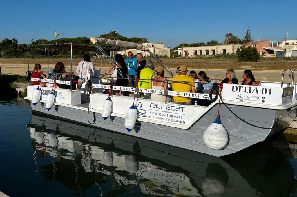 잔잔한 물과 햇살이 내리쬐는 배경의 "솔트 보트" 라는 이름의 정박된 보트에 앉아 있는 한 무리의 사람들.