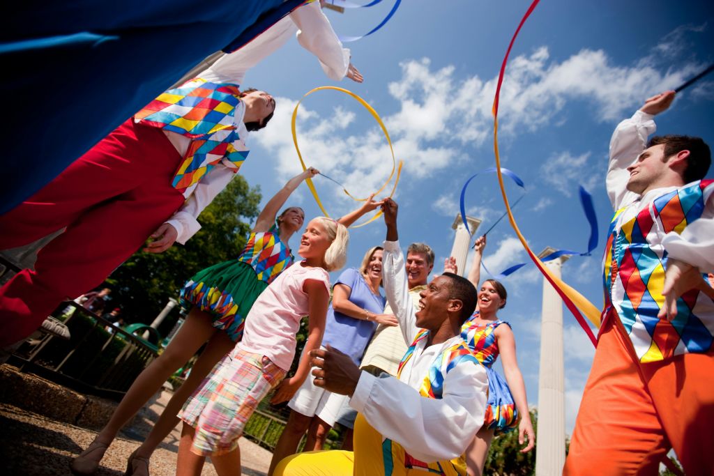화려한 의상을 입은 공연자들은 푸른 하늘 아래에서 농구대와 리본을 사용하여 야외에서 웃고 있는 가족을 즐겁게 합니다.