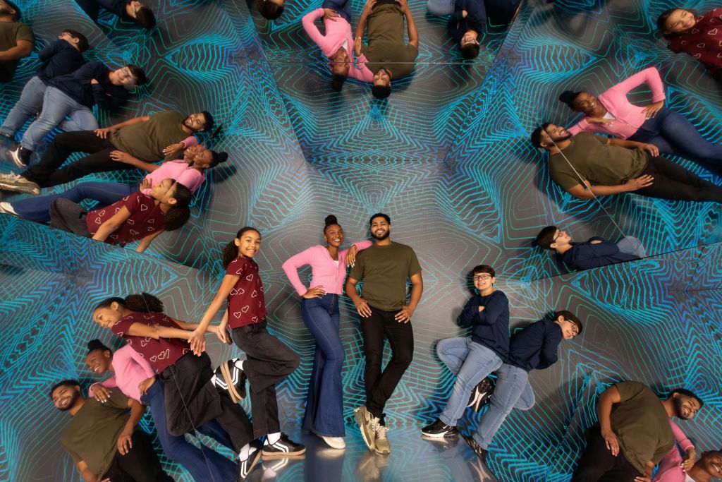 네 명의 개인이 반사 표면 앞에서 포즈를 취하며 다채로운 기하학적 배경에 여러 개의 거울 반사를 만듭니다.