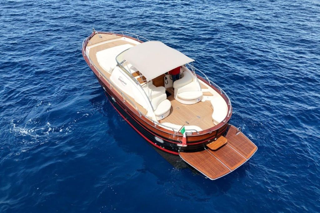 베이지색 좌석이 있는 나무 보트와 푸른 물 위를 유람하는 캐노피에는 후면 데크와 이탈리아 국기가 있습니다.