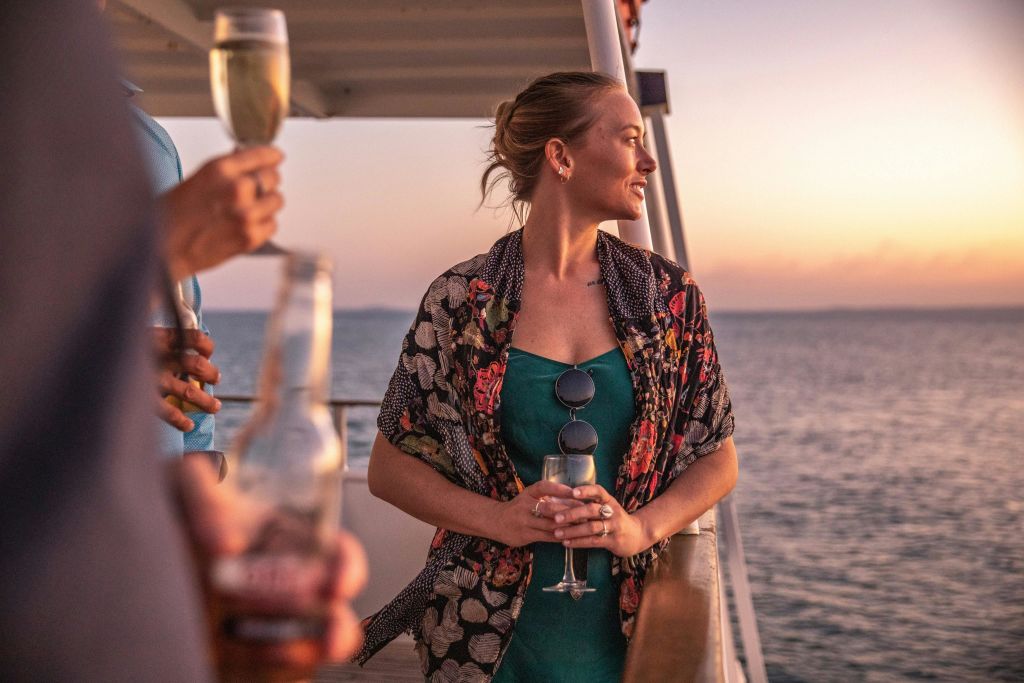 와인잔을 들고 보트에서 석양을 바라보는 여성이 초록색 드레스 위에 꽃무늬 숄을 걸치고 있습니다. 바다를 배경으로.