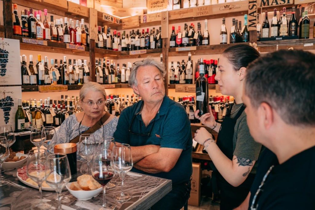 와인숍 내부에서 와인 시음회에 참석한 한 무리의 사람들로, 병을 배경으로 선반이 놓여 있습니다.