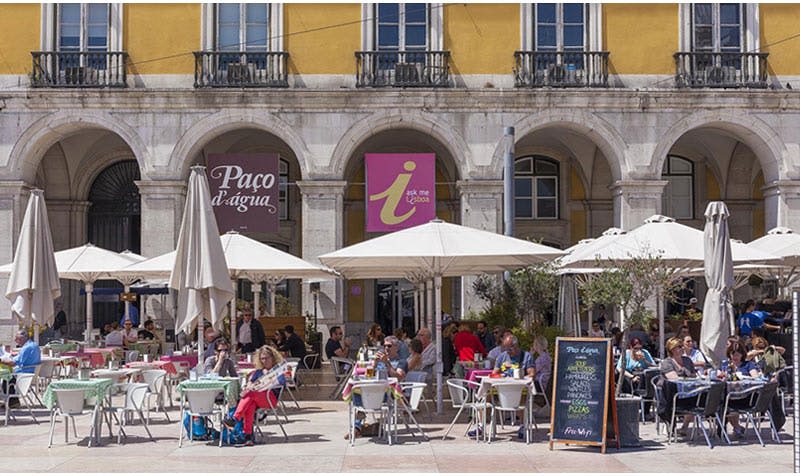 아치형 창문과 간판이 있는 노란색 건물 앞에 우산 아래 테이블에 앉아 있는 사람들이 있는 야외 카페.