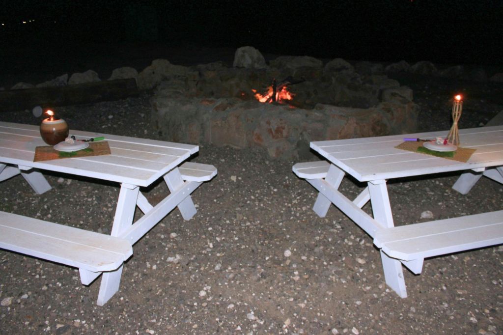 밤에 바위 바닥에 세워진 작은 불꽃이 타오르는 석조 화덕 근처에 두 개의 흰색 피크닉 테이블이 있습니다.