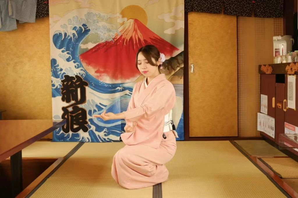 분홍색 기모노를 입은 여성이 후지산 태피스트리가 있는 방의 다다미 매트에 무릎을 꿇고 파도를 배경으로 서 있습니다.
