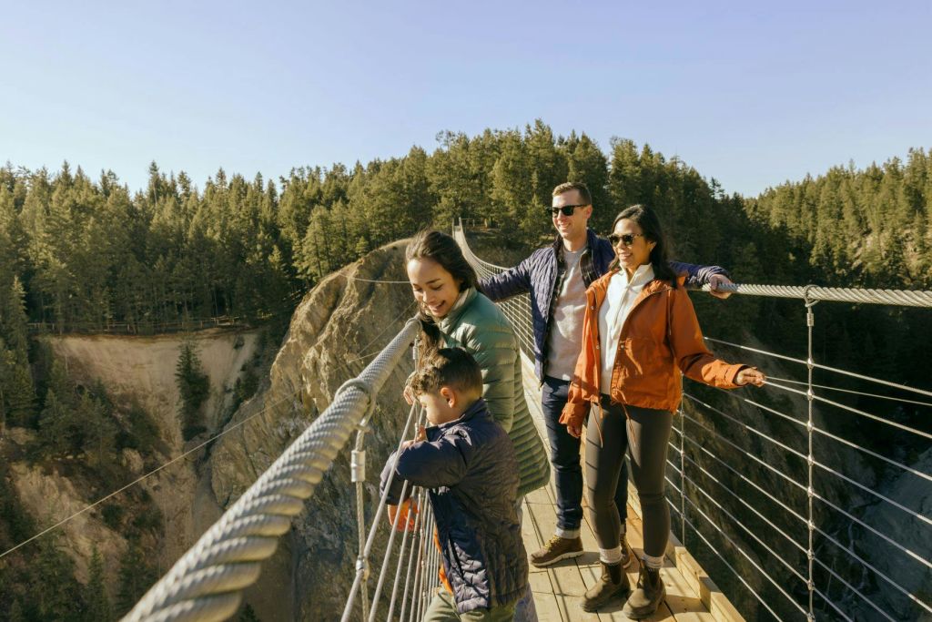 숲이 우거진 계곡 위 현수교에 4명이 서서 전망을 즐기고 있습니다. 하늘이 맑고 푸르다.