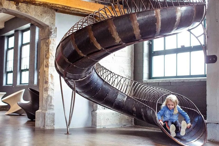 커다란 창문이 있는 인더스트리얼 스타일의 방에서 파란색 재킷을 입은 어린 아이가 실내 나선형 미끄럼틀을 타고 내려오고 있습니다.