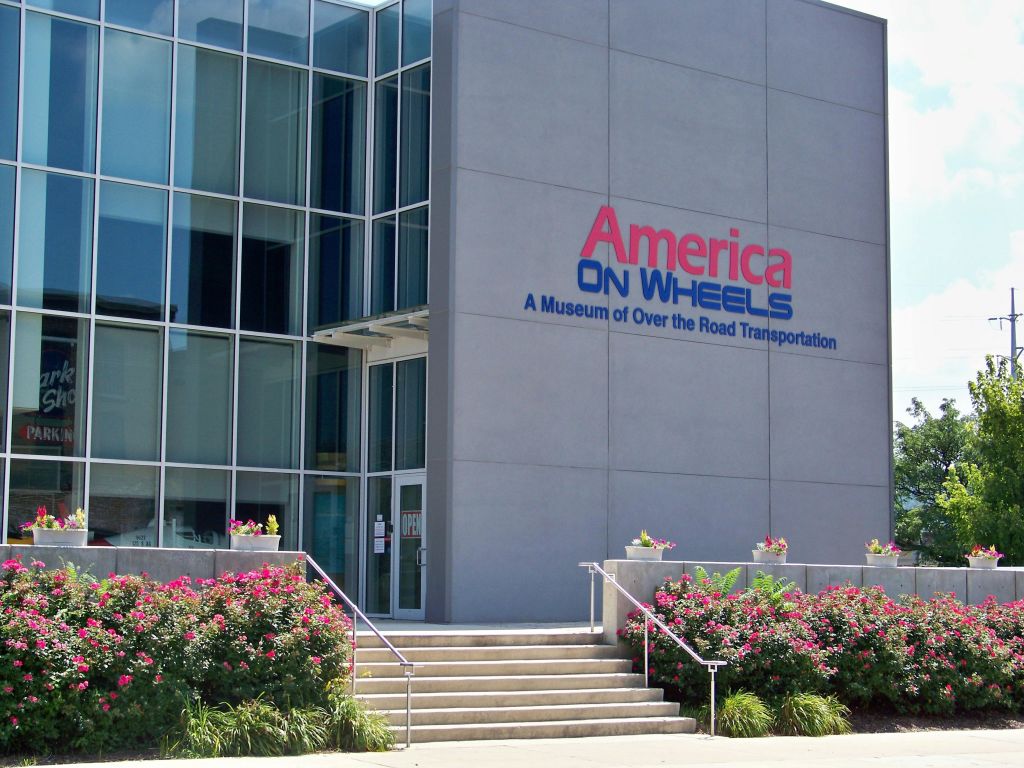 "America On Wheels: A Museum of Over the Road Transportation" 표지판, 계단, 주변 꽃이 있는 현대적인 건물 입구.