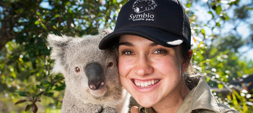 "심비오 야생동물 공원" 모자를 쓴 여성이 녹지를 배경으로 코알라 근처에 서 있습니다.