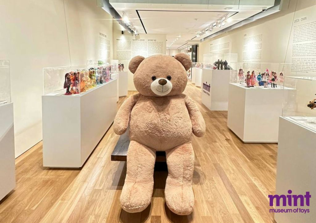 다양한 인형을 선보이는 유리 케이스가 가득한 박물관 스타일의 전시 공간에서 벤치에 앉아 있는 대형 태닝 테디 베어.