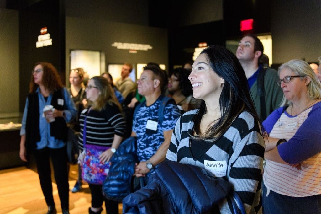 한 무리의 사람들이 실내에서 무언가를 세심하게 지켜보고 있습니다. 제니퍼라는 여성이 전경에 서서 미소를 짓고 있습니다.