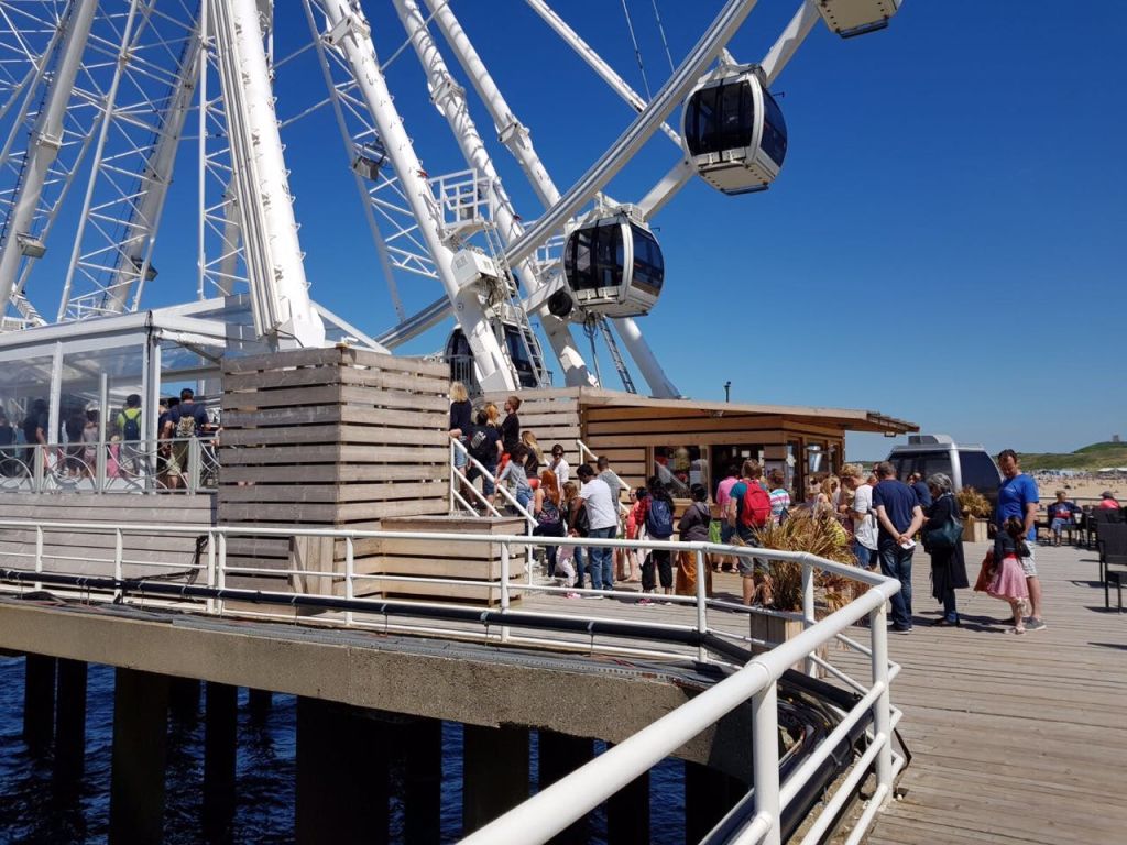 부두의 대관람차 근처에서 사람들이 줄을 서서 기다리고 있습니다. 밝고 푸른 하늘과 물이 배경입니다.