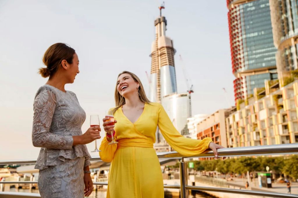 와인잔을 든 두 여성이 발코니에서 웃으며 이야기를 나누고 있습니다. 높고 공사가 진행 중인 건물이 배경입니다.