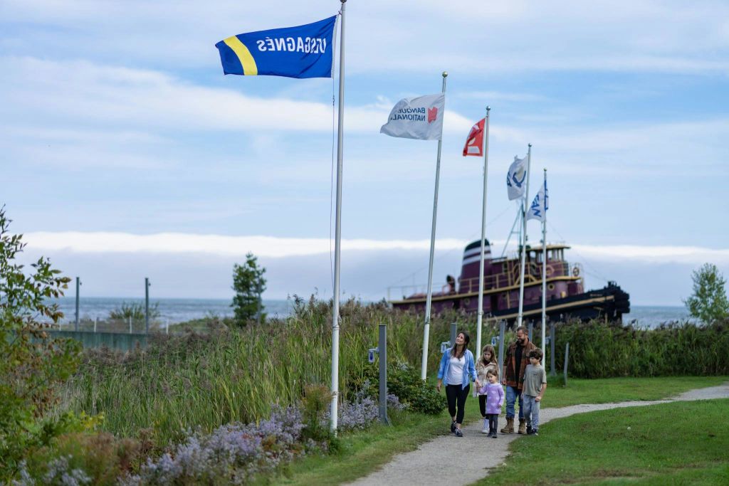 한 가족이 높은 잔디와 깃발 옆의 길을 걷고 있으며, 낡은 보트가 부분적으로 흐린 하늘 아래 배경으로 전시되어 있습니다.