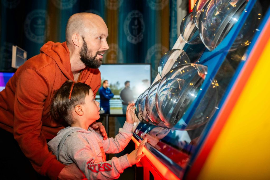 수염을 기른 남자와 어린이가 붉은 빛이 비추는 과학 전시물에서 단체 사진을 배경으로 상호 작용합니다.
