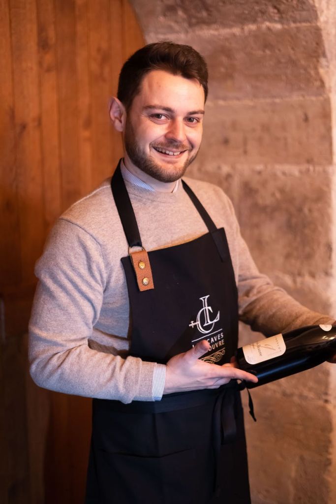 회색 스웨터를 입고 검은색 앞치마를 두른 웃는 사람이 벽돌과 나무 배경을 배경으로 실내에 서 있습니다.