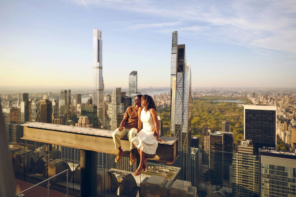 높은 건물과 커다란 공원을 배경으로 한 커플이 도시 스카이라인 위의 금속 기둥에 앉아 있습니다.