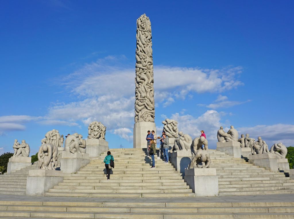 사람들은 석조 조각품으로 둘러싸인 계단을 올라가 구름이 흩어져 있는 푸른 하늘을 배경으로 높은 석상으로 이어집니다.