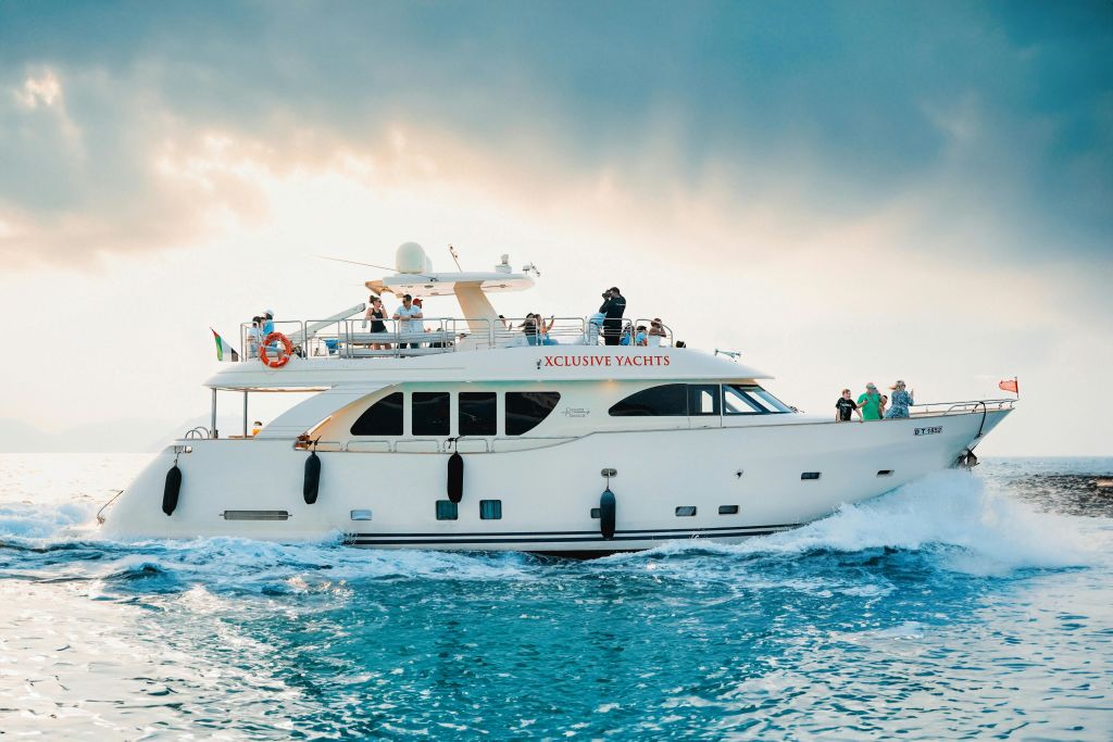 흐린 하늘 아래 갑판에 사람들이 있는 푸른 물 위의 하얀 고급 요트. 요트 옆면에는 "전용 요트" 라고 쓰여 있습니다.