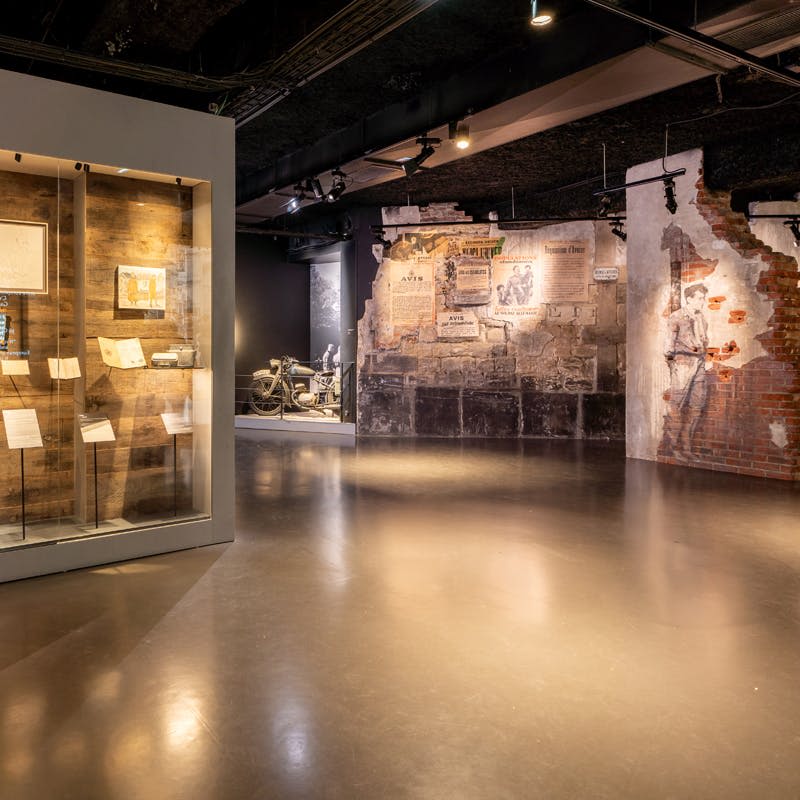 문서와 유물이 전시된 전시 케이스와 벽화와 빈티지 오토바이가 있는 주변 지역이 있는 박물관 전시관입니다.