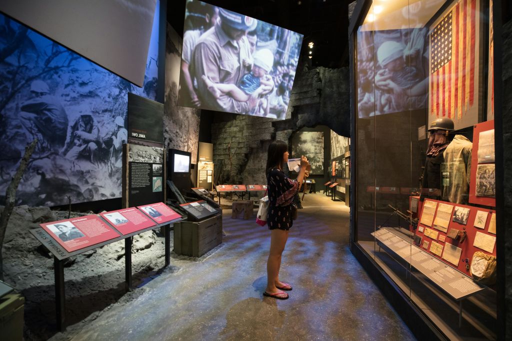 한 여성이 전시 영상을 보여주는 대형 스크린 아래에서 역사적인 이미지와 텍스트가 담긴 박물관 전시물을 촬영하고 있습니다.