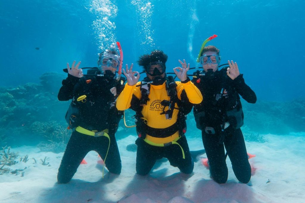 수중에서 잠수복과 마스크를 착용한 세 명의 스쿠버 다이버가 맑고 푸른 물과 모래가 깔린 바다를 배경으로 무난한 손팻말을 만듭니다.