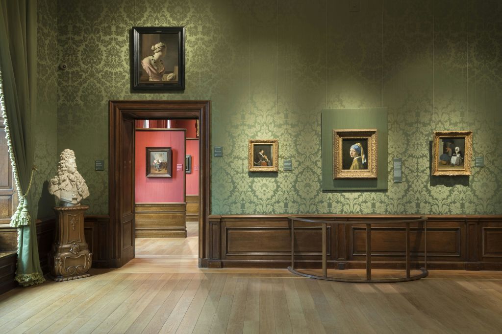 녹색 벽지, 나무 바닥, 액자에 담긴 그림이 있는 박물관 방. 출입구를 지나면 붉은 벽이 있는 다른 갤러리가 나옵니다.