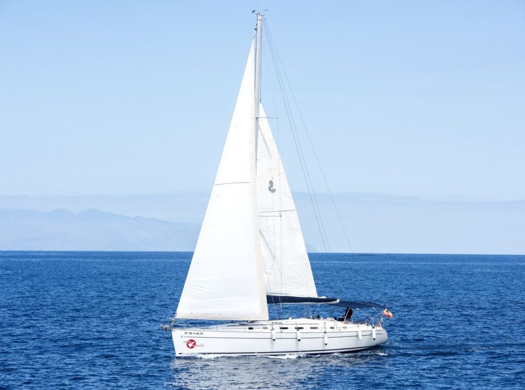 커다란 돛을 단 하얀 요트가 저 멀리 산이 펼쳐진 맑은 하늘 아래 잔잔하고 푸른 바다를 미끄러지고 있습니다.