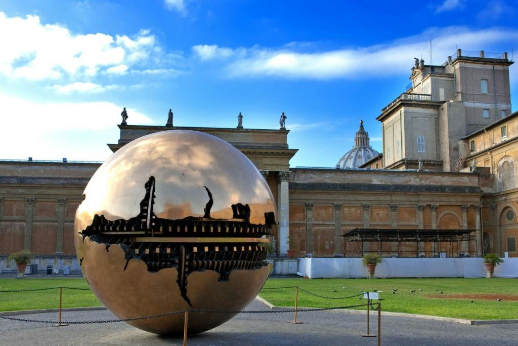 화창한 날에 고전 건축물 근처 야외에 내부 구조를 갖춘 구의 커다란 청동 조각상이 전시되어 있습니다.