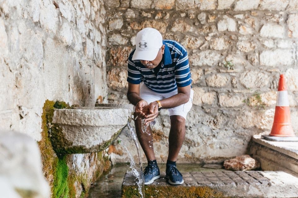 줄무늬 셔츠와 흰색 마개를 쓴 사람이 오래된 돌 분수에서 질감이 살아있는 돌담을 배경으로 물을 마시고 있습니다.