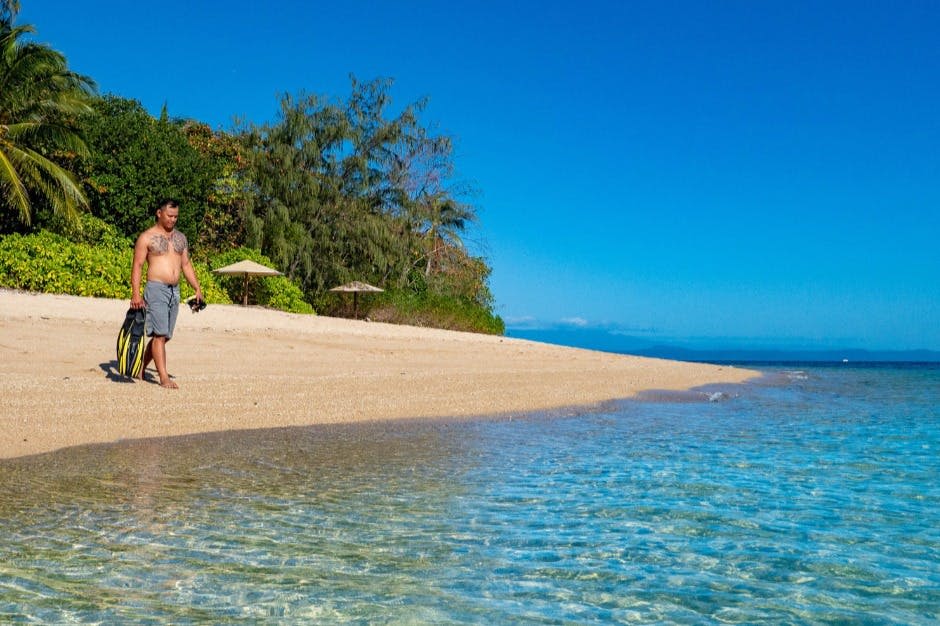 맑고 푸른 물이 흐르는 모래사장에 한 남성이 서 있습니다. 맑은 하늘 아래 배경으로 비치 파라솔과 나무가 보입니다.
