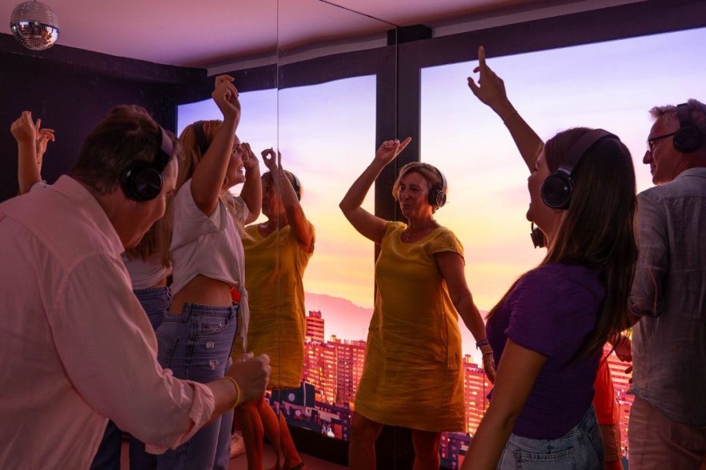 해 질 녘 도시 풍경을 보여주는 커다란 창문이 있는 방에서 헤드폰을 착용한 사람들이 열정적으로 춤을 추고 있습니다.