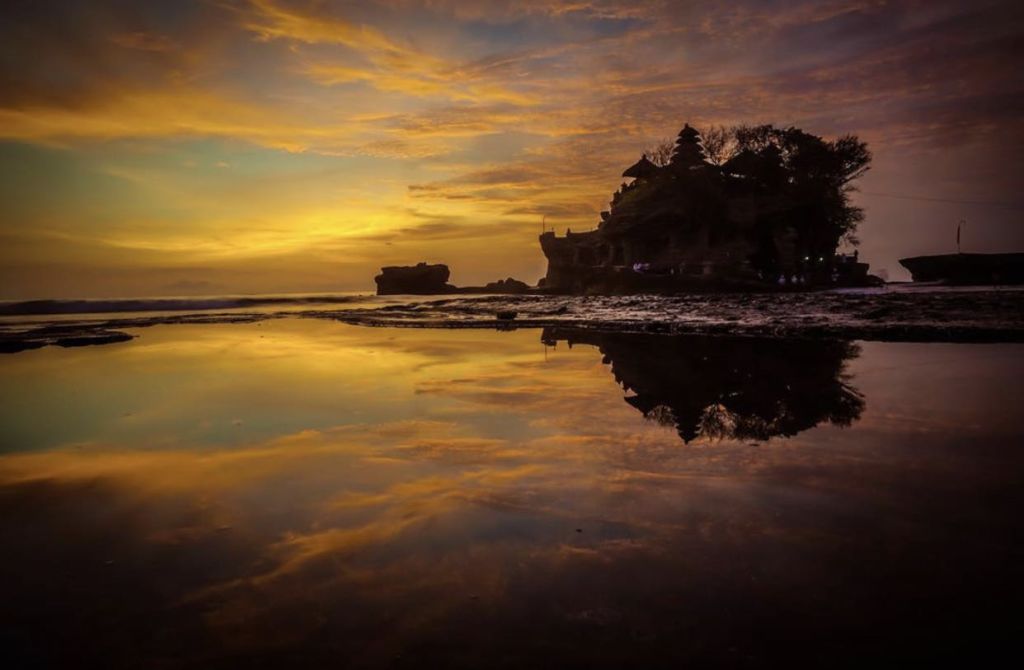 잔잔한 물속에 비친 하늘을 배경으로 사원이 있는 바위 해안선 너머로 지는 석양.