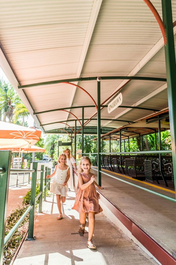 세 명의 어린 소녀가 "엔더톰" 이라고 적힌 표지판 옆 그늘진 산책로에서 즐겁게 달립니다. 배경에는 녹지와 빈 벤치가 포함됩니다.