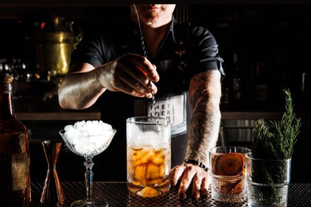 바텐더가 믹싱 글래스에 얼음이 담긴 음료를 저어줍니다. 근처에 얼음 한 잔과 오렌지 슬라이스로 장식된 칵테일이 있습니다.