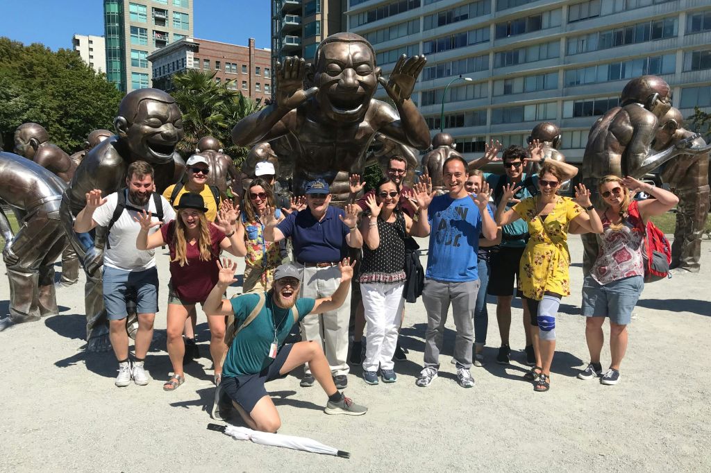 화창한 날 야외 도심에서 청동으로 만든 웃는 조각상 앞에서 포즈를 취하고 있는 한 무리의 사람들.