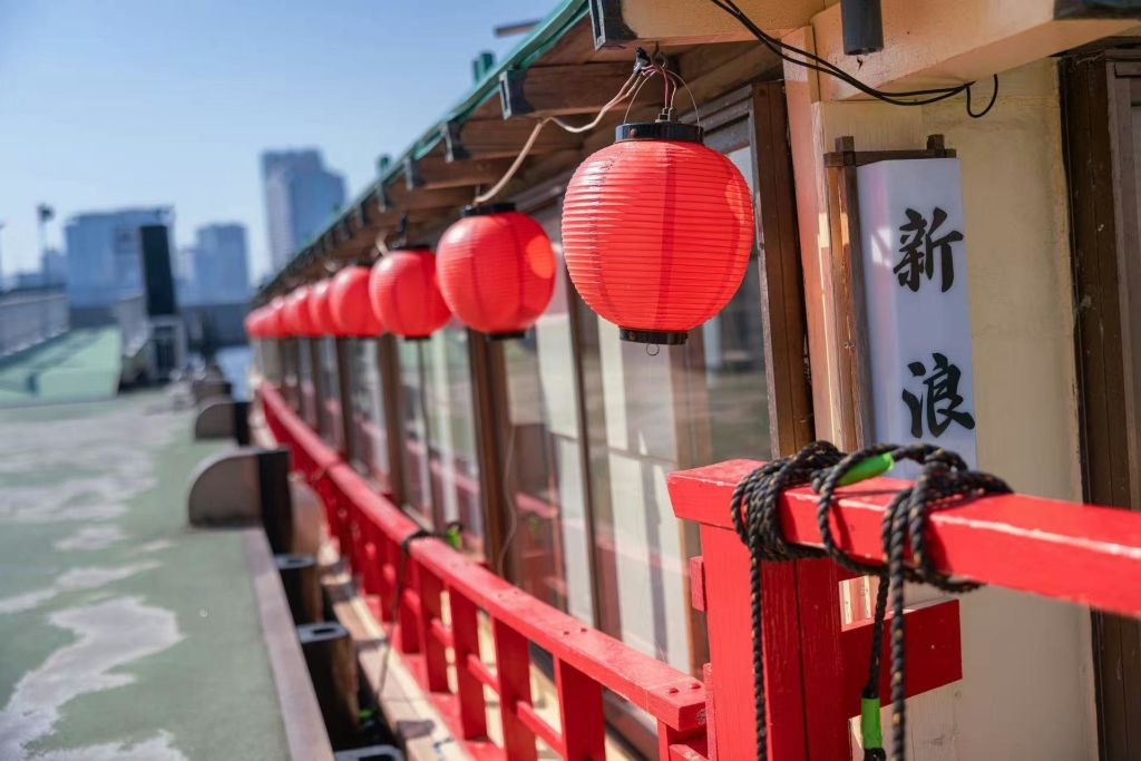 도시 풍경과 푸른 하늘을 배경으로 빨간 난간이 있는 전통 건물 외부에 빨간 등불이 줄지어 걸려 있습니다.