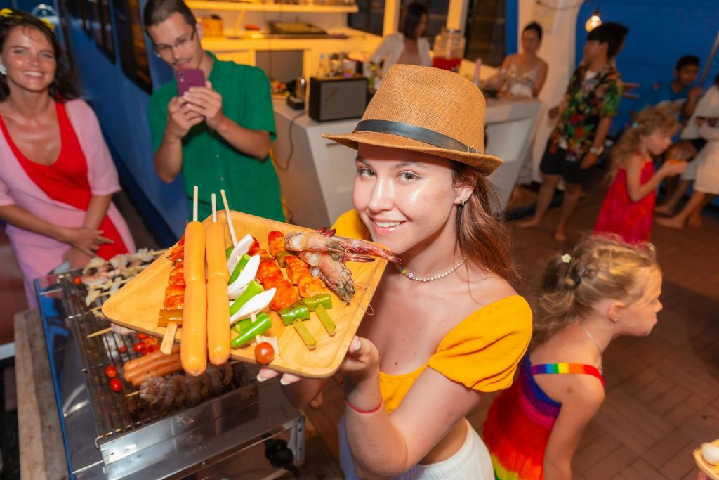 활기찬 실내 바비큐 파티에서 고기, 야채, 소시지가 담긴 꼬치 트레이를 들고 있는 모자를 쓴 여성.