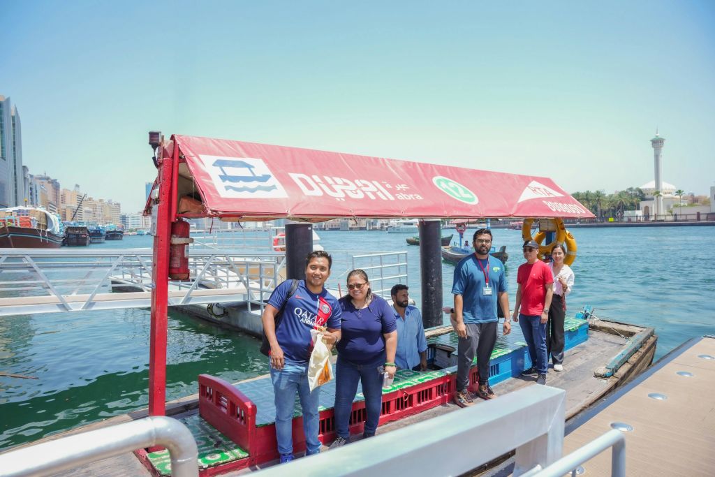 화창한 날 보트와 물을 배경으로 붉은 캐노피 아래 선착장에 서 있는 6명의 사람들.