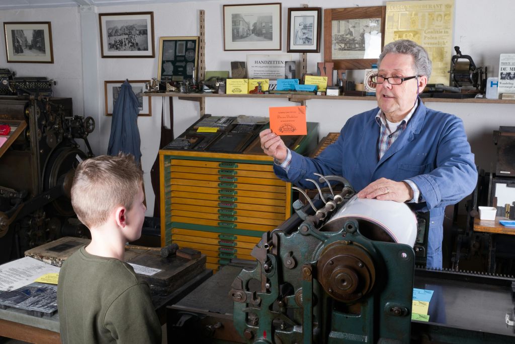 파란색 재킷을 입은 한 노인이 인쇄 장비와 액자 그림으로 가득 찬 방에서 어린 소년에게 주황색 카드를 보여줍니다.