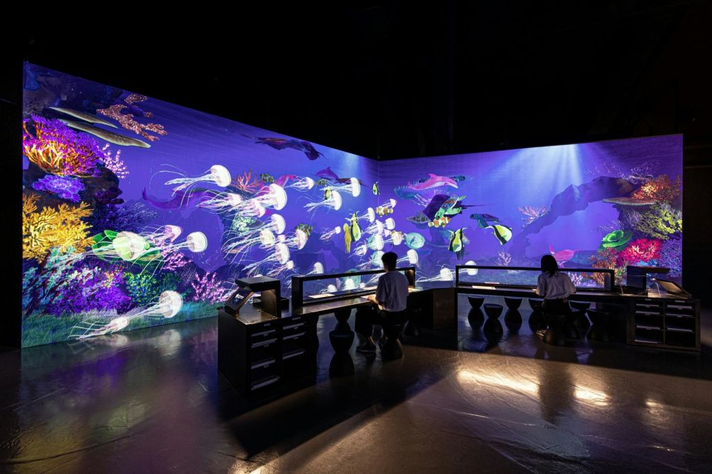 해파리, 화려한 물고기 등 해양 생물이 전시된 대형 수족관 스크린이 있는 방의 책상에 두 사람이 앉아 있습니다.