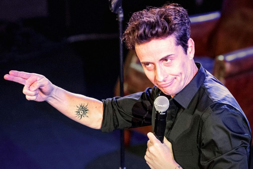 검은 셔츠를 입고 마이크를 들고 무대에서 팔에 문신을 새긴 채 말하고 있는 사람.