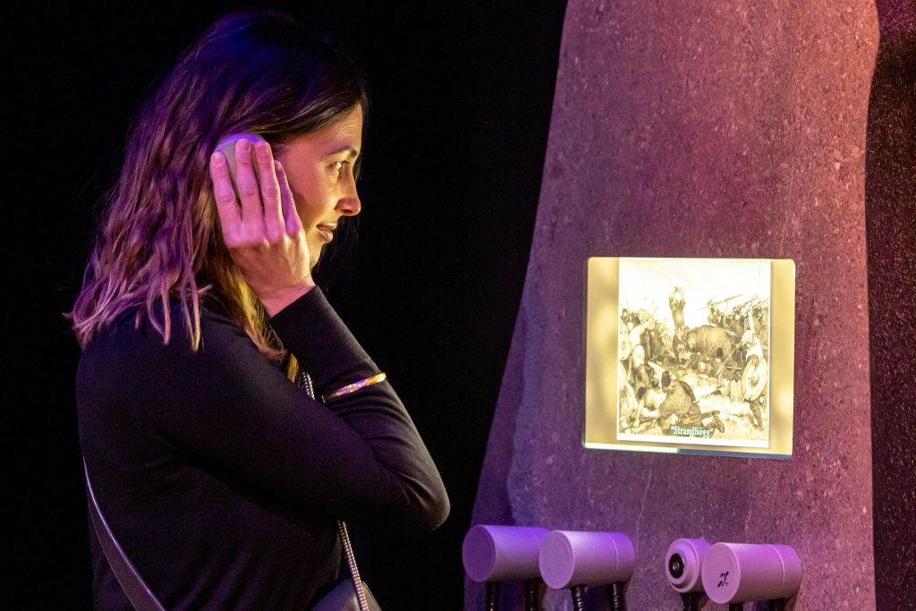 한 여성이 조명이 어두운 조명 패널 근처에 서서 기기를 귀에 대고 경청하고 있습니다.